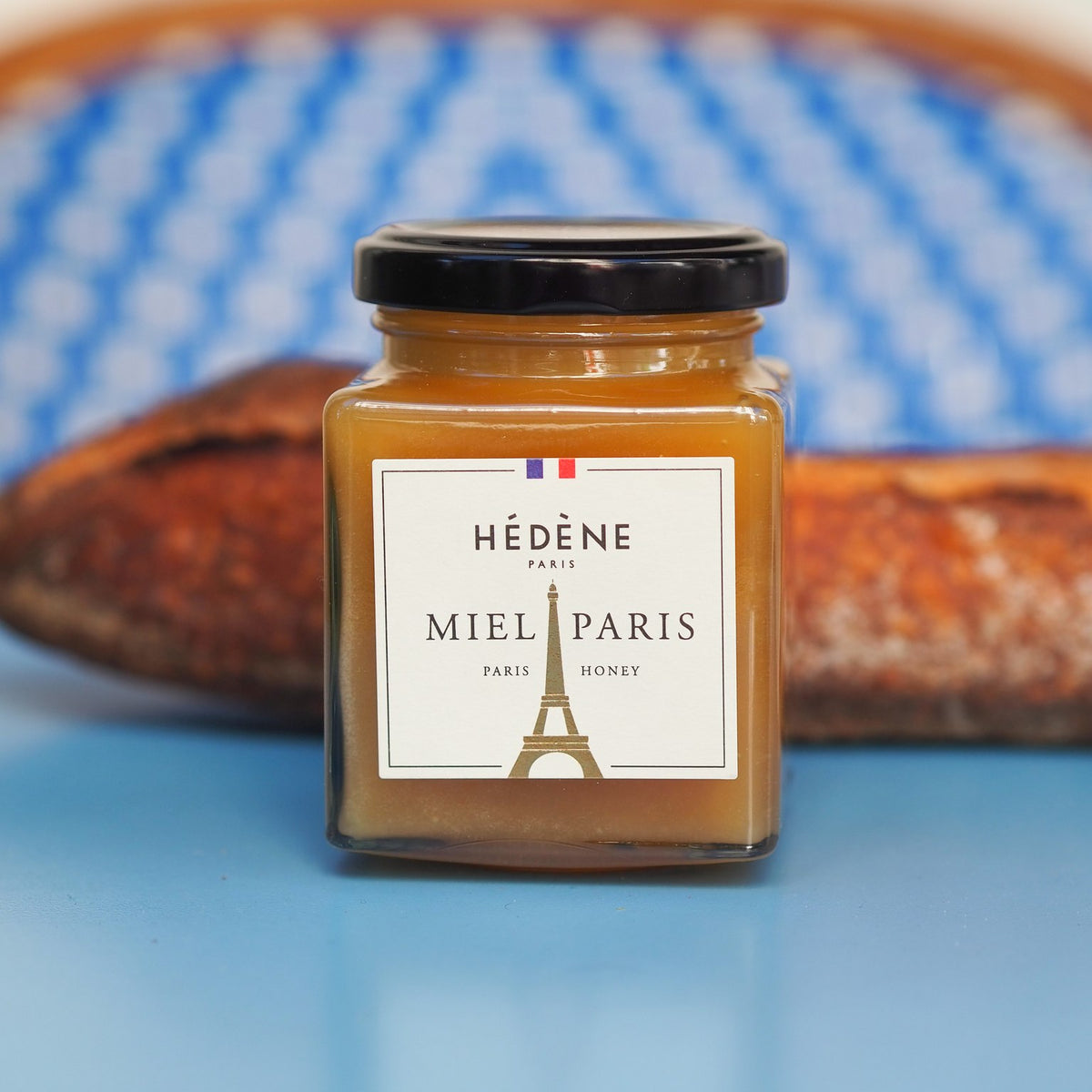 Pot de Miel de Paris Hédène à l’étiquette dorée ornée de la tour Eiffel, devant une baguette et une chaise de bistrot